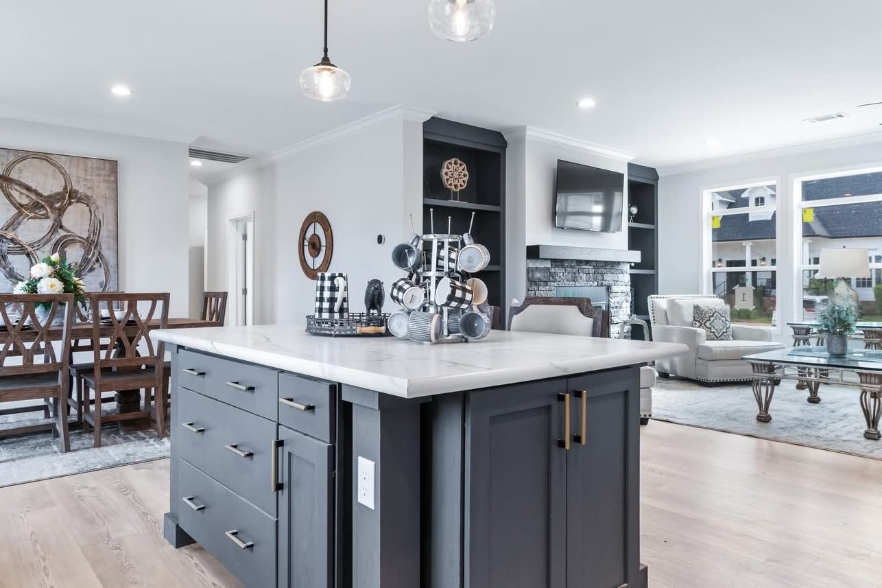 Modern, airy kitchen and living area with a gray island, pendant lights, dining table, and cozy seating by a fireplace. Large windows provide natural light.