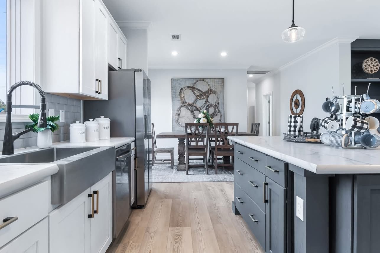 Modern kitchen-dining space with white cabinets, gray island, and stainless-steel sink. Dining table with flowers in background. Bright, tidy ambiance.