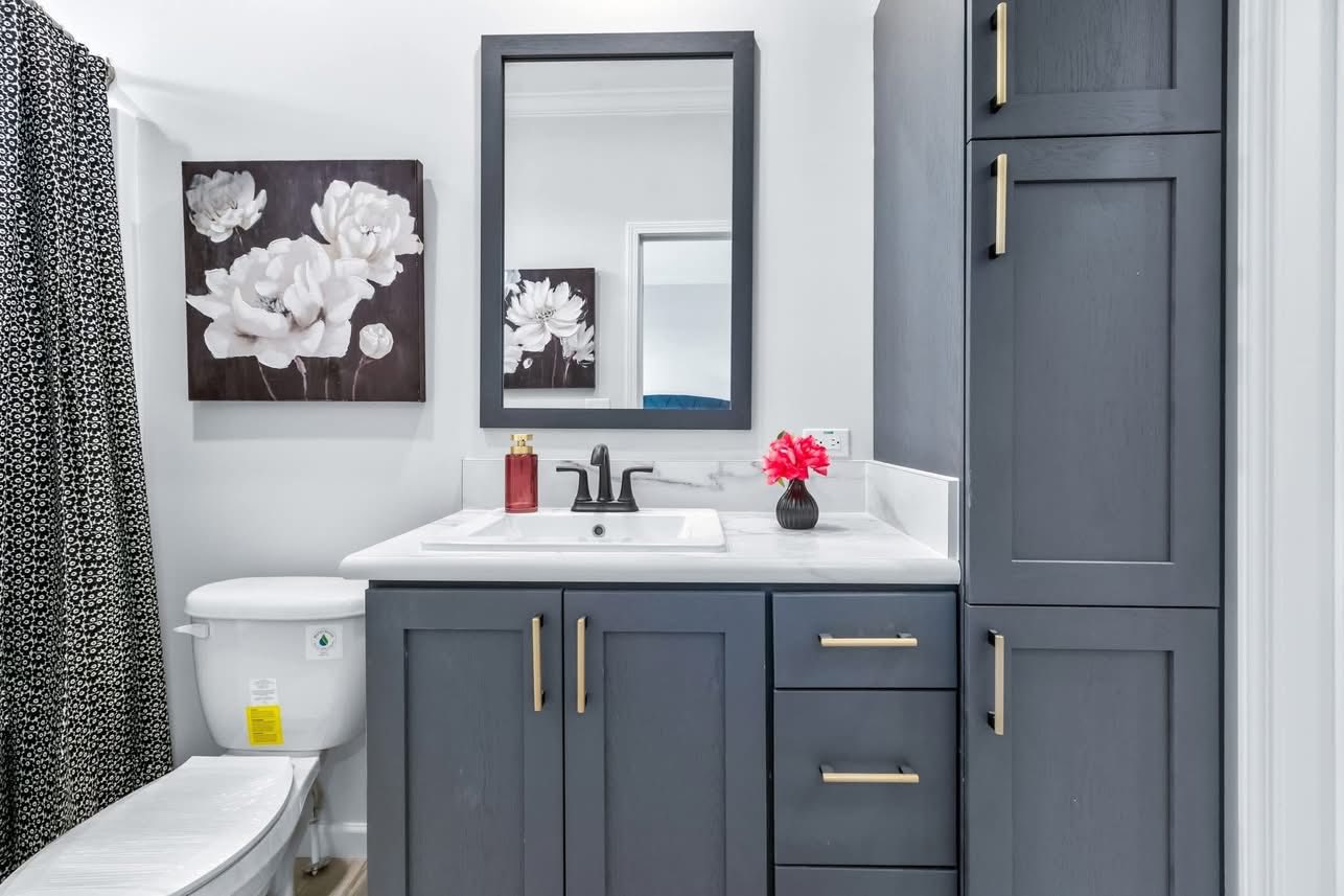 Modern bathroom with gray cabinets and gold handles. White flowers in a dark frame hang above the toilet. Black faucet and red flower on the sink.