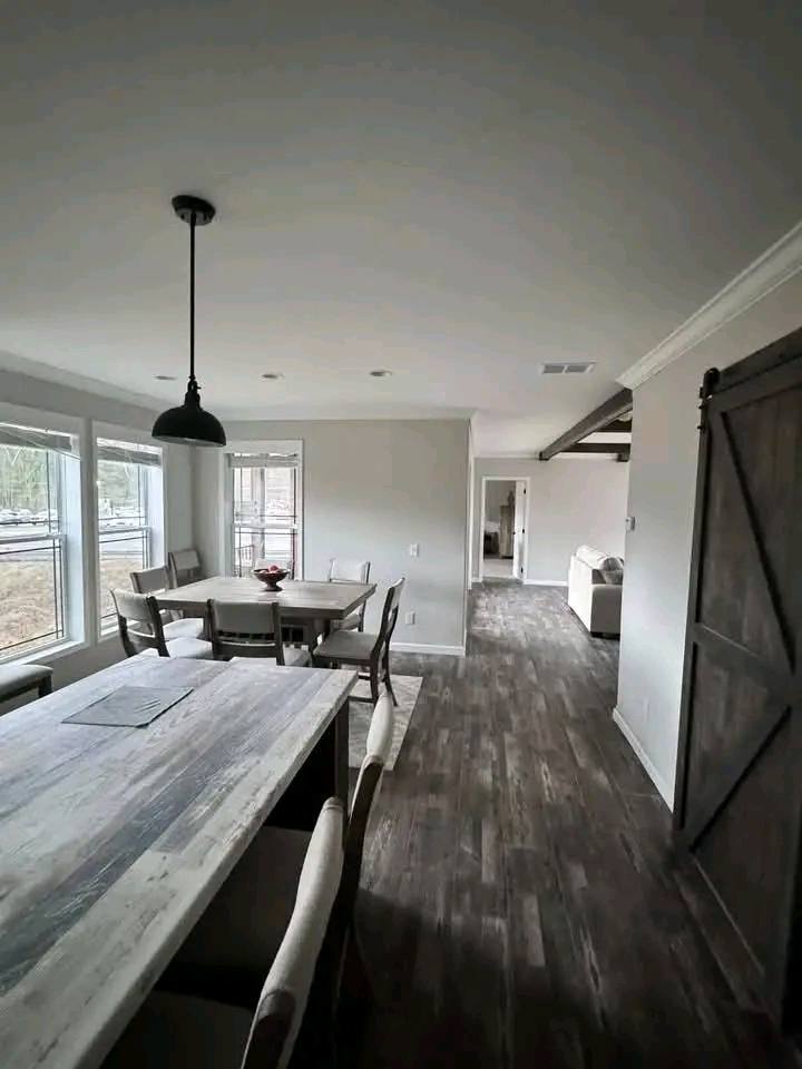 Modern dining room with a rustic wooden table and chairs, large windows, and a black pendant light. Hallway leads to a living area with wood flooring.
