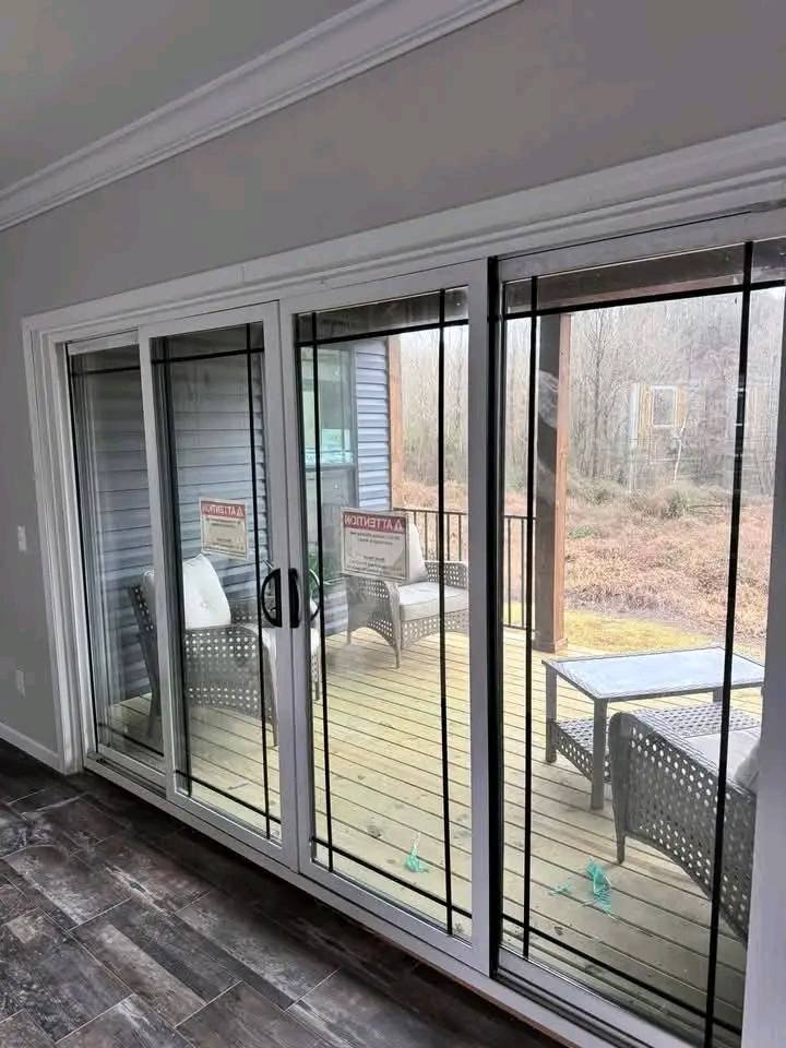 View through glass sliding doors opens to a wooden deck with wicker chairs and table, accented by gray siding and misty forest beyond. Calm ambiance.