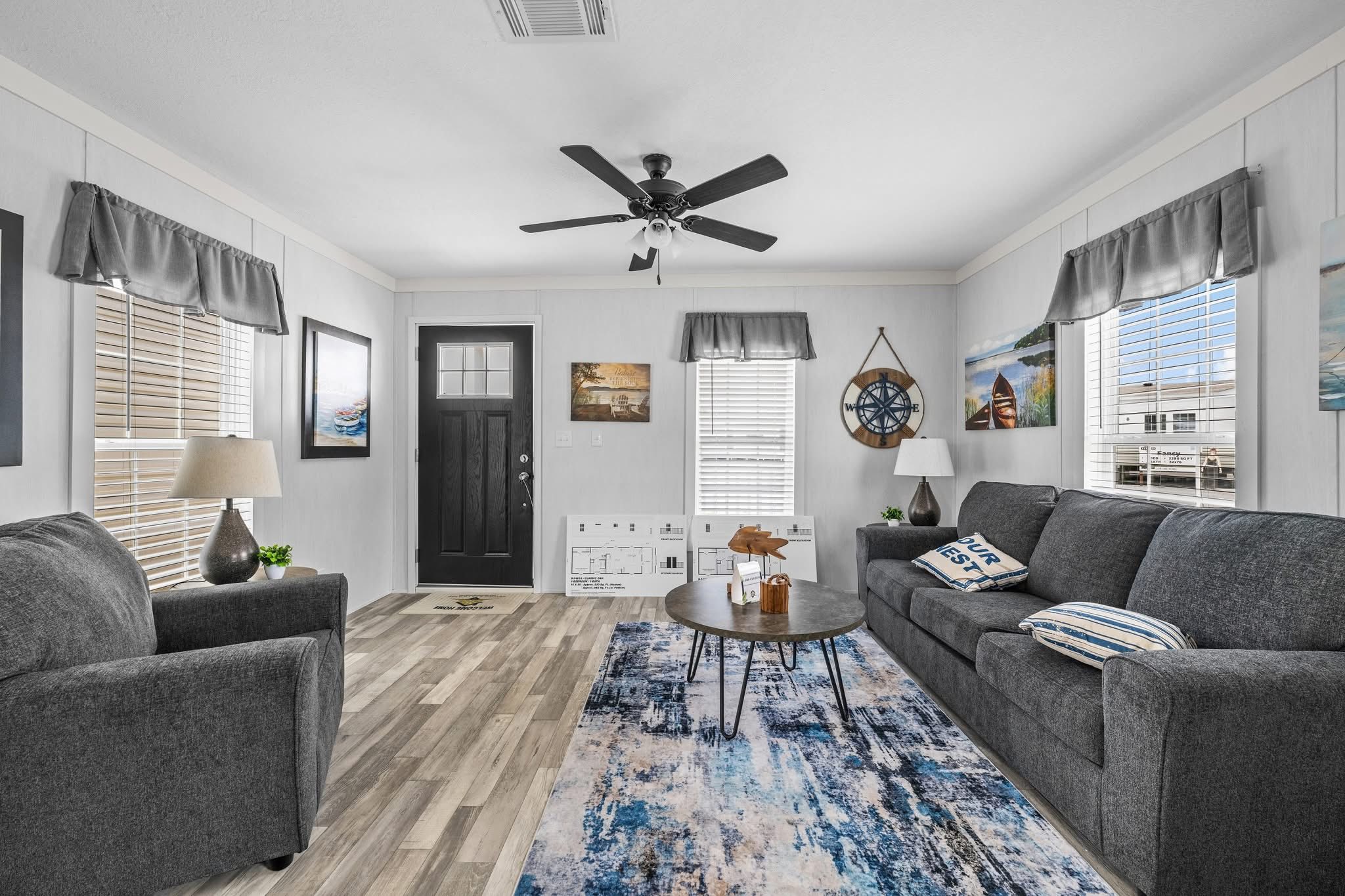 Cozy living room with gray sofas, blue and white rug, and round coffee table. Nautical decor and soft lighting create a welcoming, calm atmosphere.