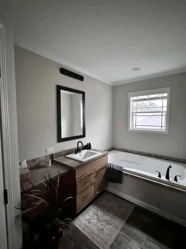 A modern bathroom with a sleek design. A wooden vanity with a white sink and black faucet is next to a bathtub. A large mirror hangs above, and a potted plant adds warmth.