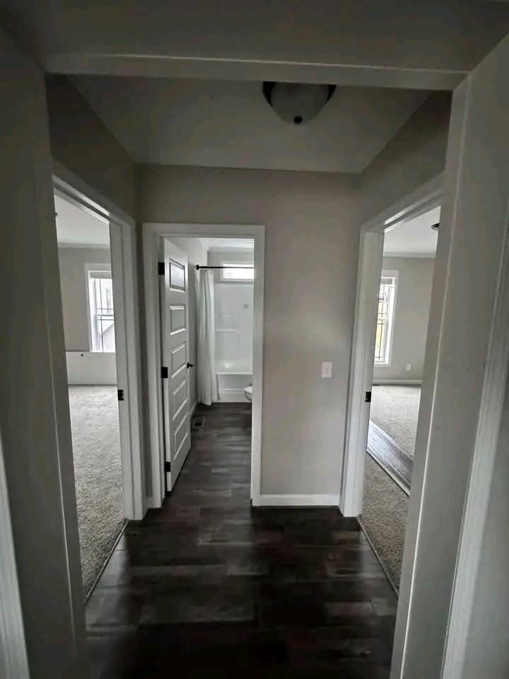 Hallway with dark wood floor, leading to three rooms. Left and right doors open to carpeted rooms with windows; center door reveals a bathroom with white fixtures.