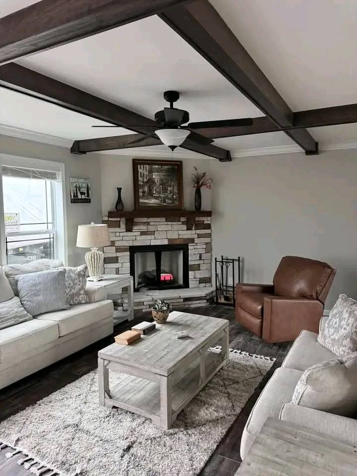 Cozy living room with beige sofas, a brown armchair, and a stone fireplace. Ceiling beams, modern fan, and soft lighting create a warm, inviting atmosphere.