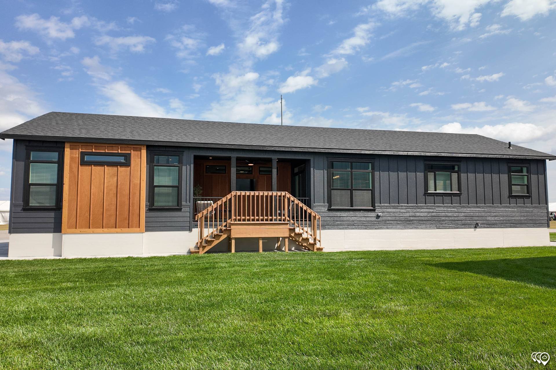 Single-story modern house with gray and wooden exterior, large windows, and a small porch with stairs, set against a blue sky and green lawn.