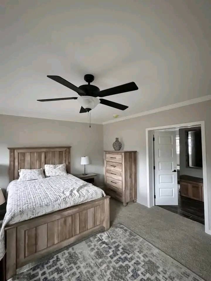 Cozy bedroom with a wooden bed, patterned bedding, matching dresser, and nightstand. A ceiling fan and soft lighting create a warm ambiance.