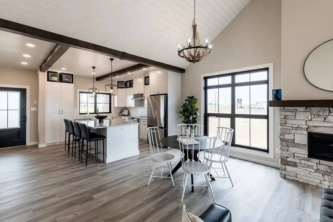 Spacious open-concept kitchen and dining area with vaulted ceilings. Features a white island, black chairs, stone fireplace, large windows, and a chandelier.