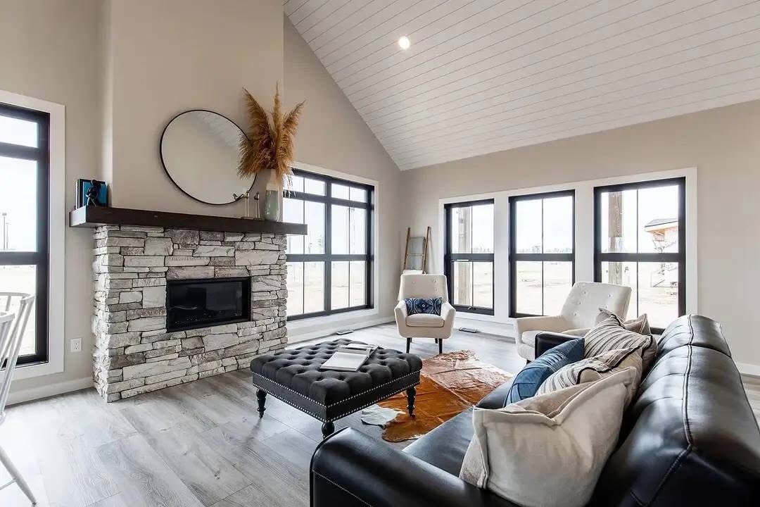 A modern living room with a vaulted ceiling, stone fireplace, and large windows. Includes a black leather couch, plush armchairs, and a tufted ottoman. Cozy and bright.
