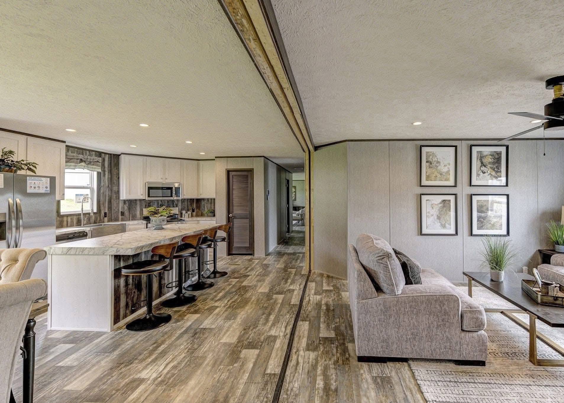 Modern open-plan interior featuring a kitchen with a marble island and bar stools. Living area has a gray sofa, coffee table, plants, and framed art.