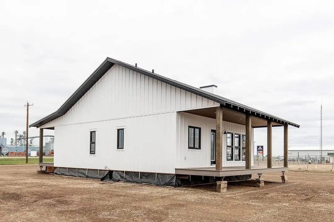 A white modular home with a sloped metal roof and front porch on wooden stilts. It sits on a dirt lot with an overcast sky, conveying a minimalist, rural feel.