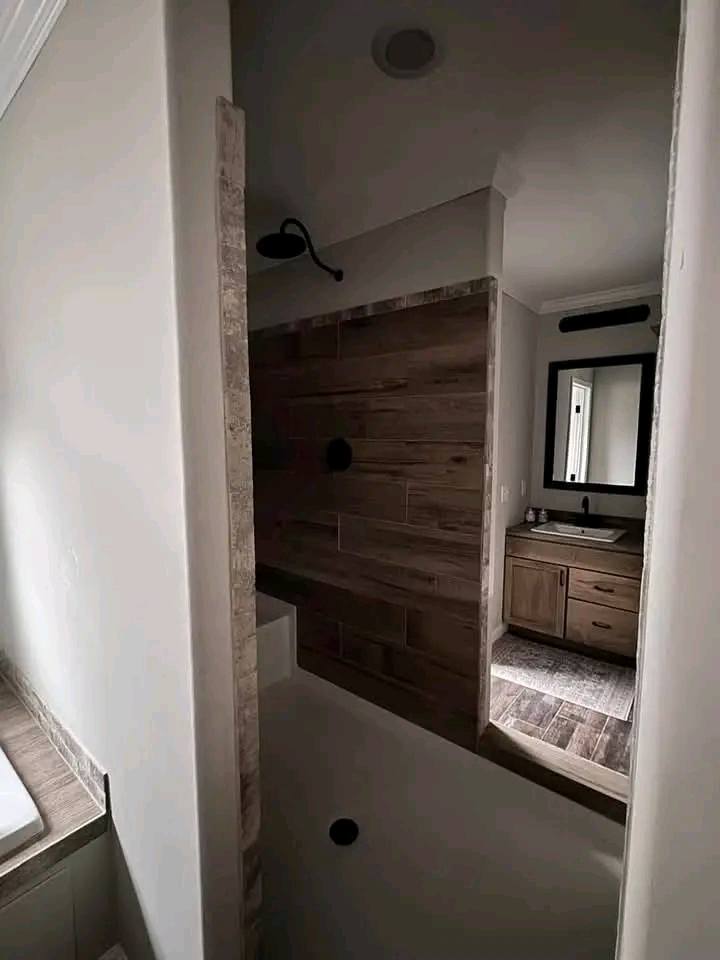Dark, modern bathroom with wooden walls and flooring. A showerhead is mounted on the left. A vanity with a mirror is in the background, creating a cozy feel.