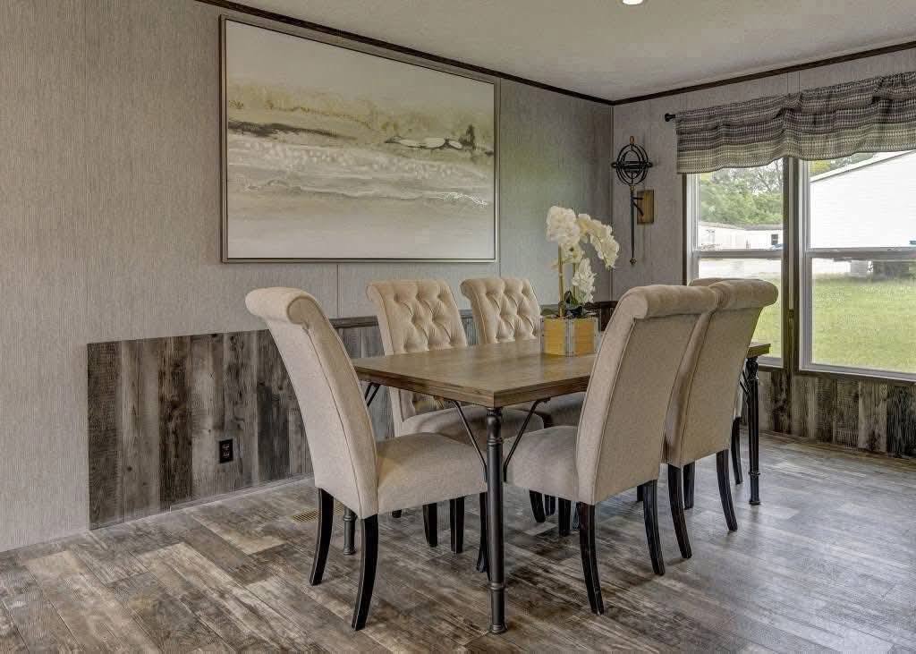 A dining room with a wooden table and eight beige upholstered chairs. A large abstract painting hangs on the wall. Soft, natural light fills the space.
