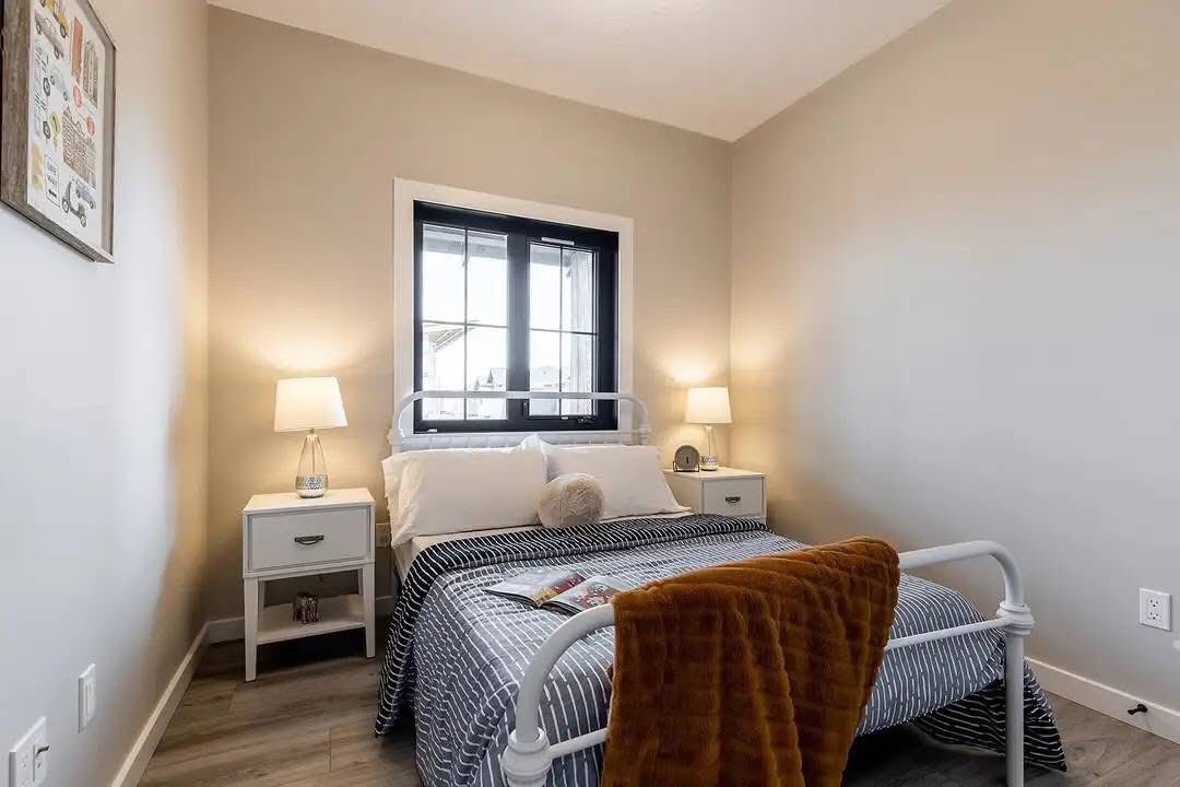 Cozy bedroom with a metal-framed bed, striped bedding, and white pillows. A soft brown throw drapes over the footboard. Warm lighting from bedside lamps on white nightstands creates an inviting atmosphere.