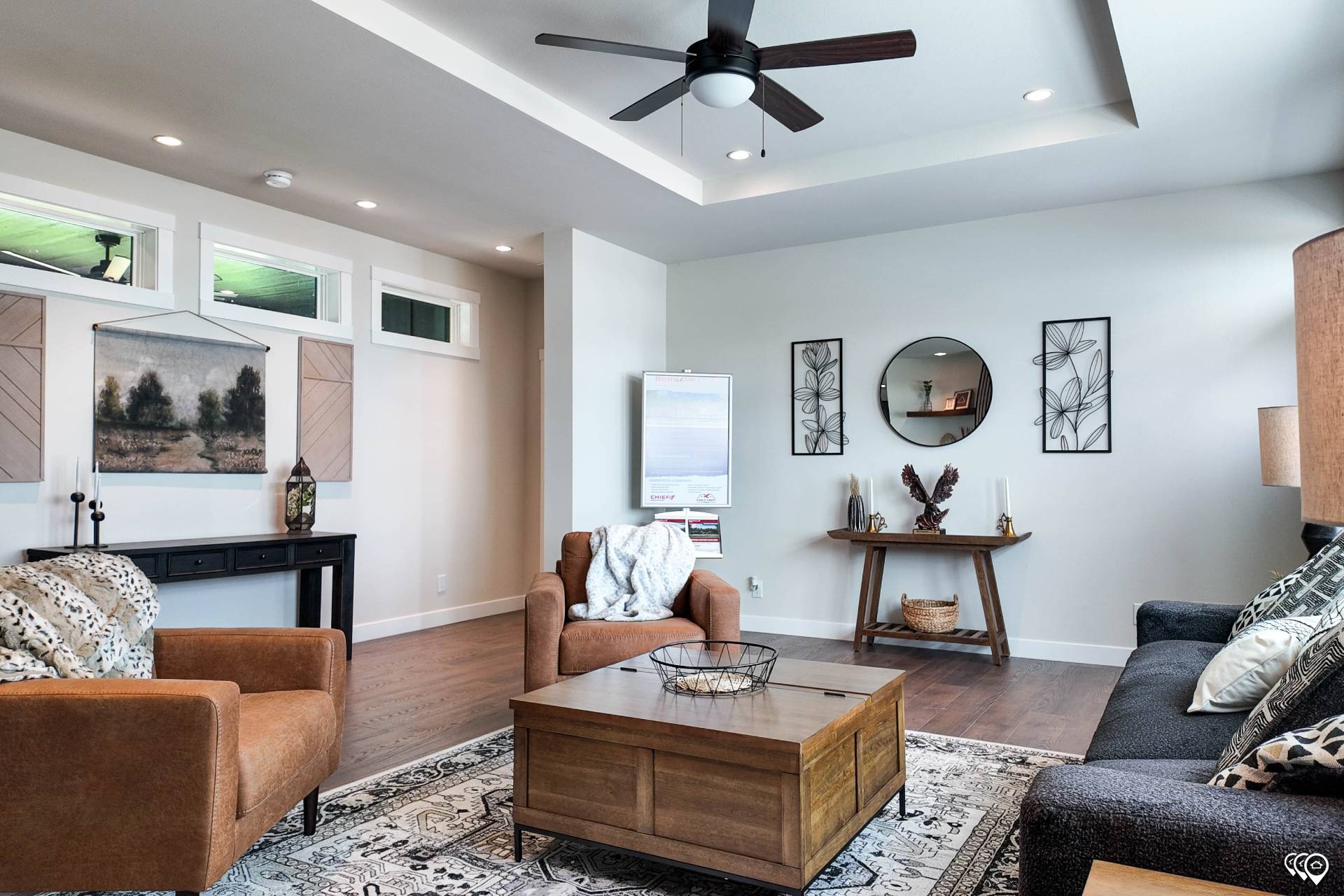 Modern living room with brown armchairs, a black sofa, and a wooden coffee table on a patterned rug. Wall art and a ceiling fan add elegance.