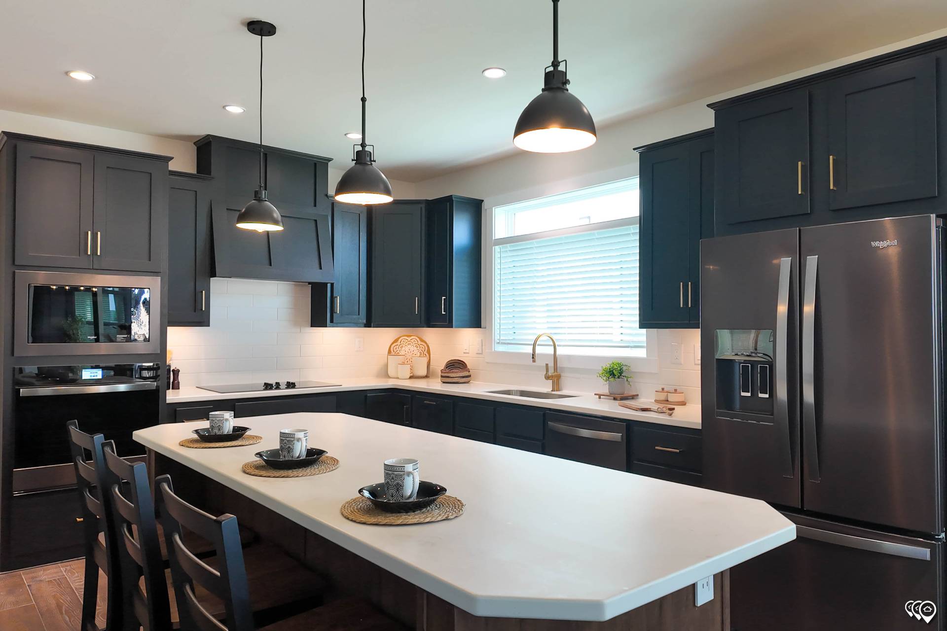 Modern kitchen with dark cabinets, white countertops, and a large island with four chairs. Three pendant lights hang above, creating a cozy atmosphere.