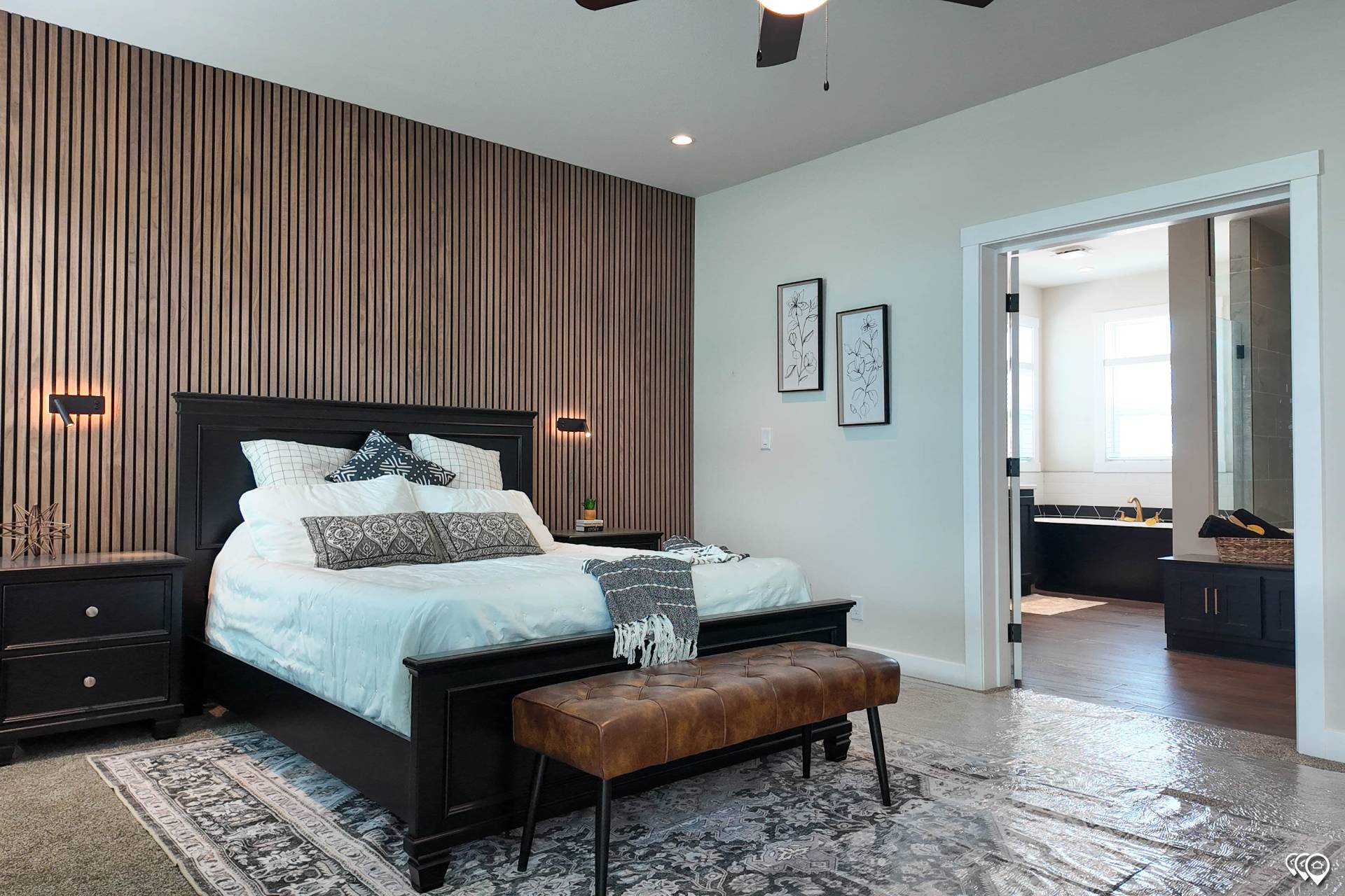 Modern bedroom with a cozy, inviting atmosphere. Features a dark bed frame against a wooden slat wall, soft lighting, a patterned area rug, and an adjacent open bathroom.