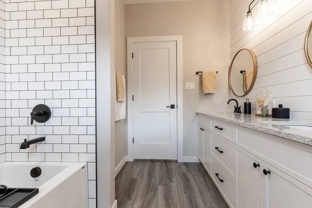 Modern bathroom with white subway tile shower, black fixtures, light wood floor, dual round mirrors, and white vanity with granite countertop.