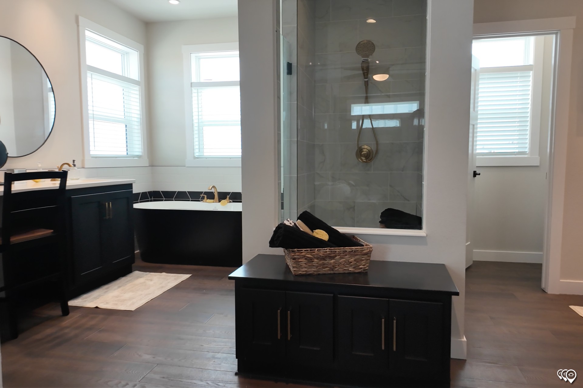 Modern bathroom with dark wood cabinets, gold fixtures, a round mirror, a black bathtub, and a glass shower. Bright and elegant ambiance.