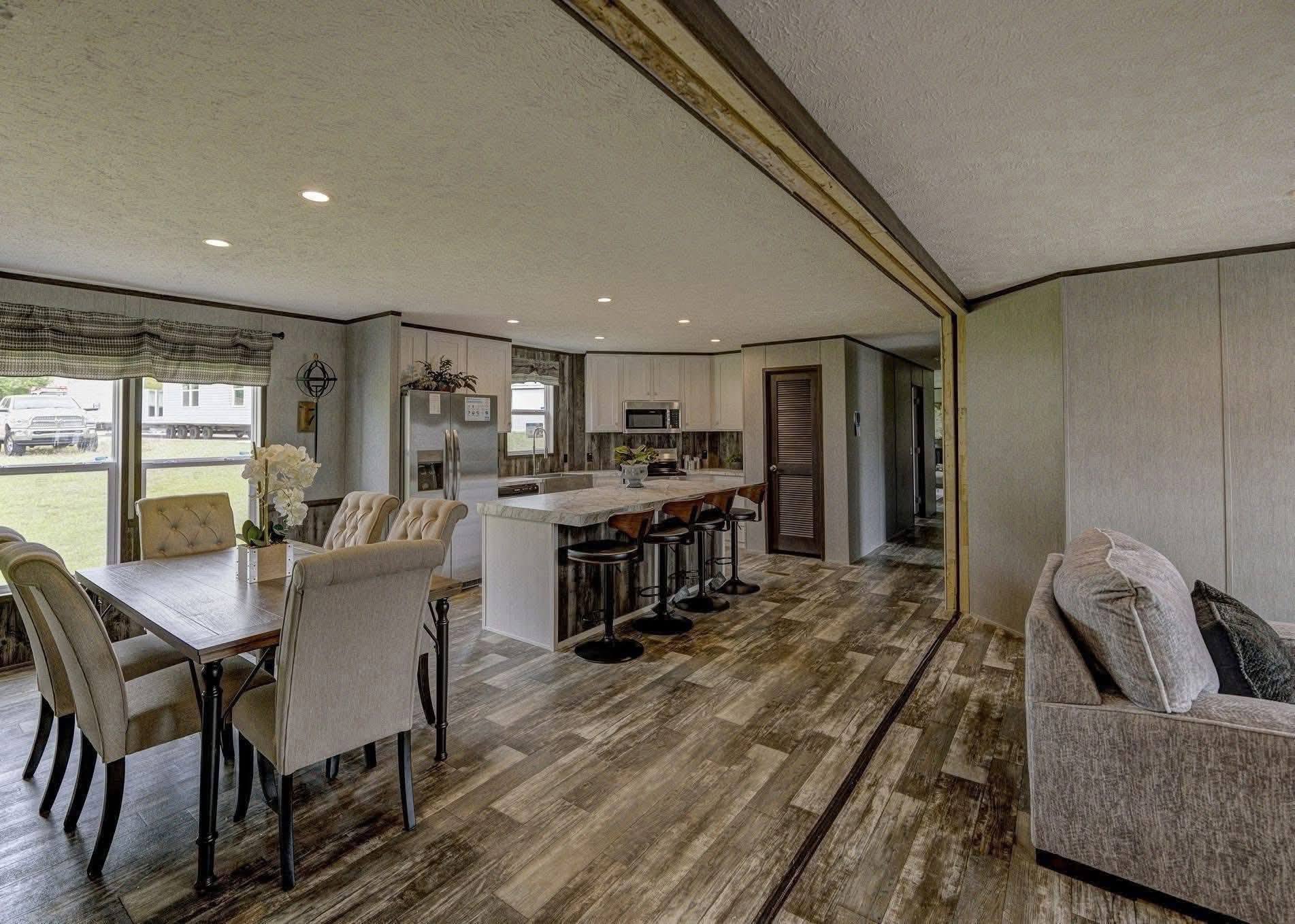 Open-concept living space with a wooden floor, featuring a dining table with beige chairs and a kitchen with bar stools. Bright and inviting ambiance.