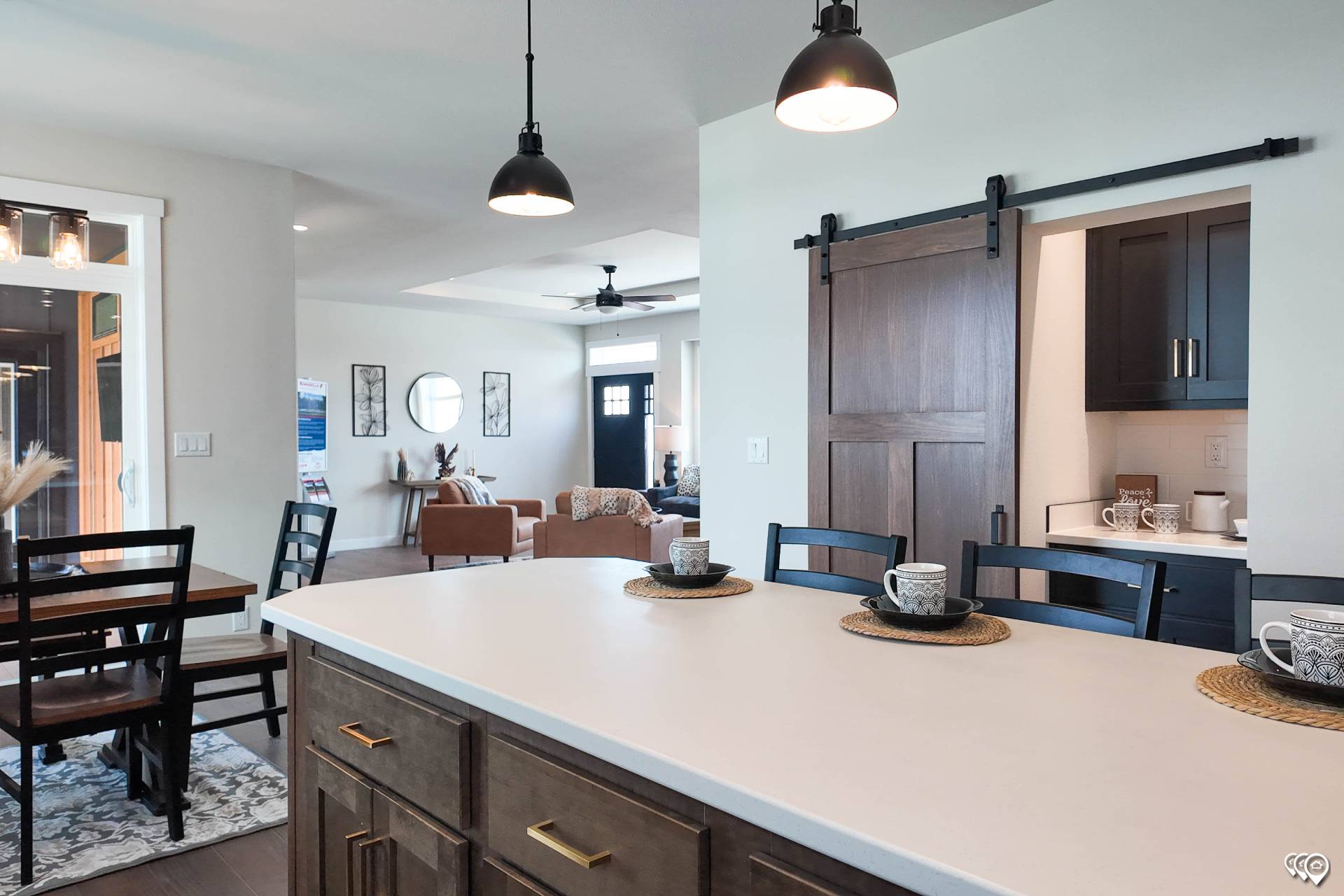 Modern kitchen and dining area with a large island, pendant lights, and bar stools. A barn door leads to a cozy living room with neutral tones. Warm and inviting ambiance.