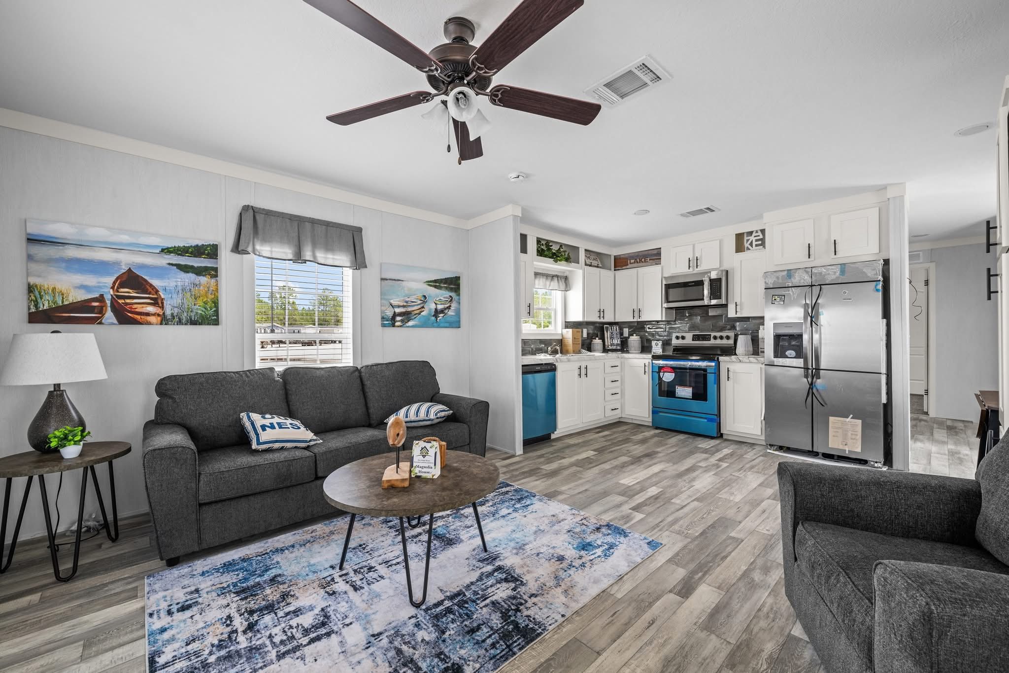Cozy living room and kitchen with gray sofas, round coffee table, nautical-themed decor, stainless steel appliances, and a ceiling fan. Bright and inviting.