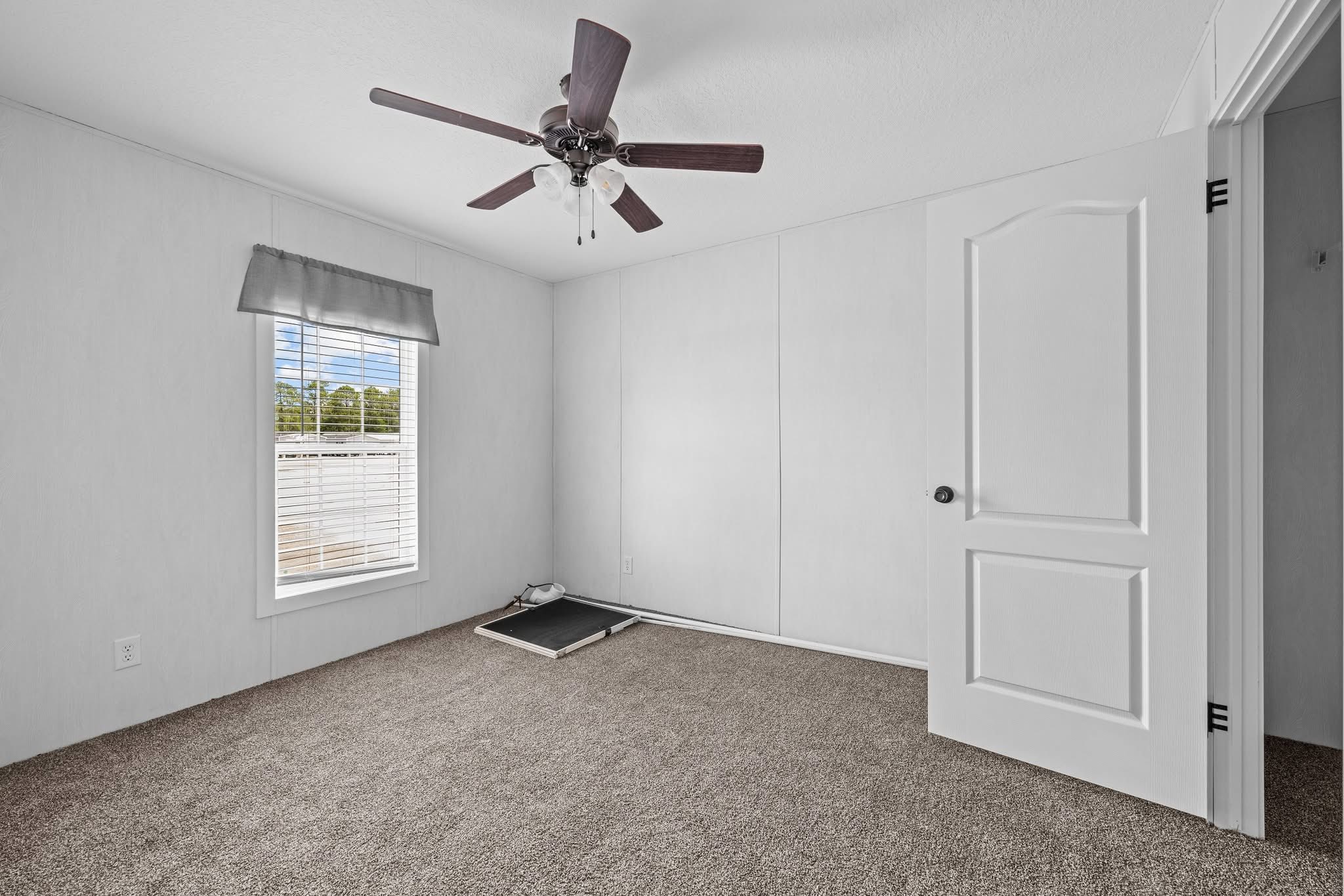 Empty room with beige carpet and white walls. A ceiling fan with dark wood blades is on the ceiling. A window with white blinds and a grey valance lets in natural light. A treadmill is folded against the wall, and an open door is visible on the right.