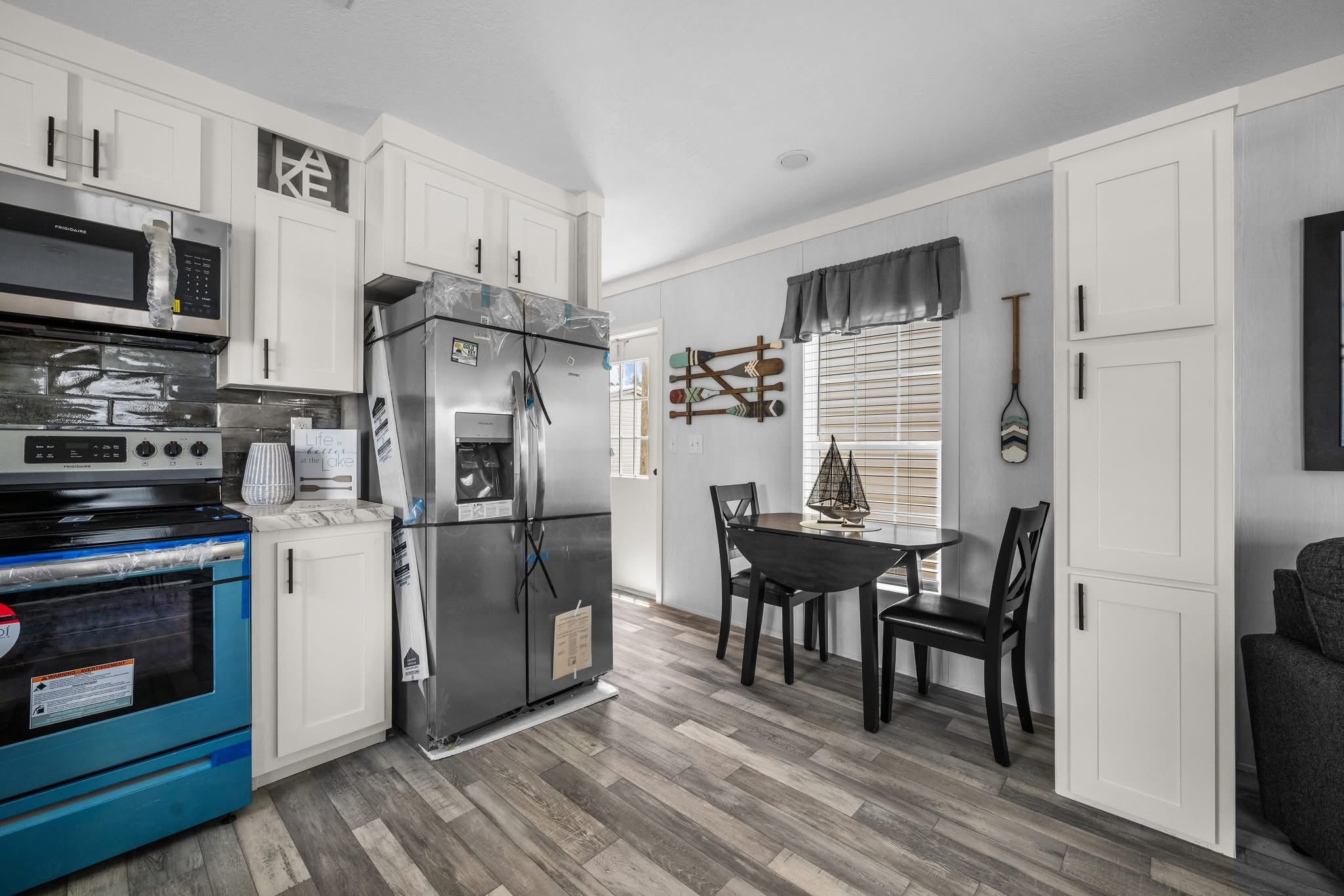 Modern kitchen with white cabinets, stainless steel appliances, and wood laminate flooring. Small dining area with two chairs by a bright window.