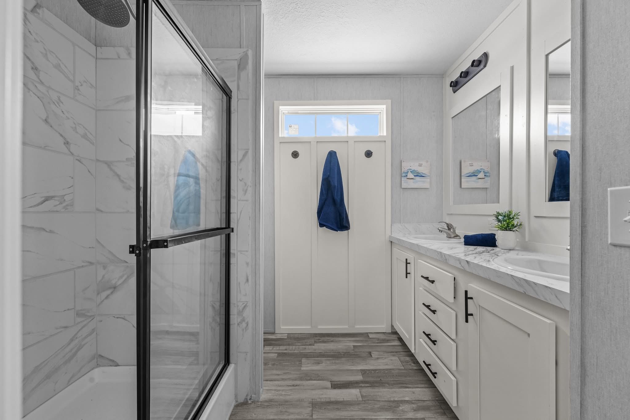 Modern bathroom featuring a glass-enclosed shower with marble tiles, a double vanity with white cabinets and a marble countertop, and a blue towel hanging near a window.
