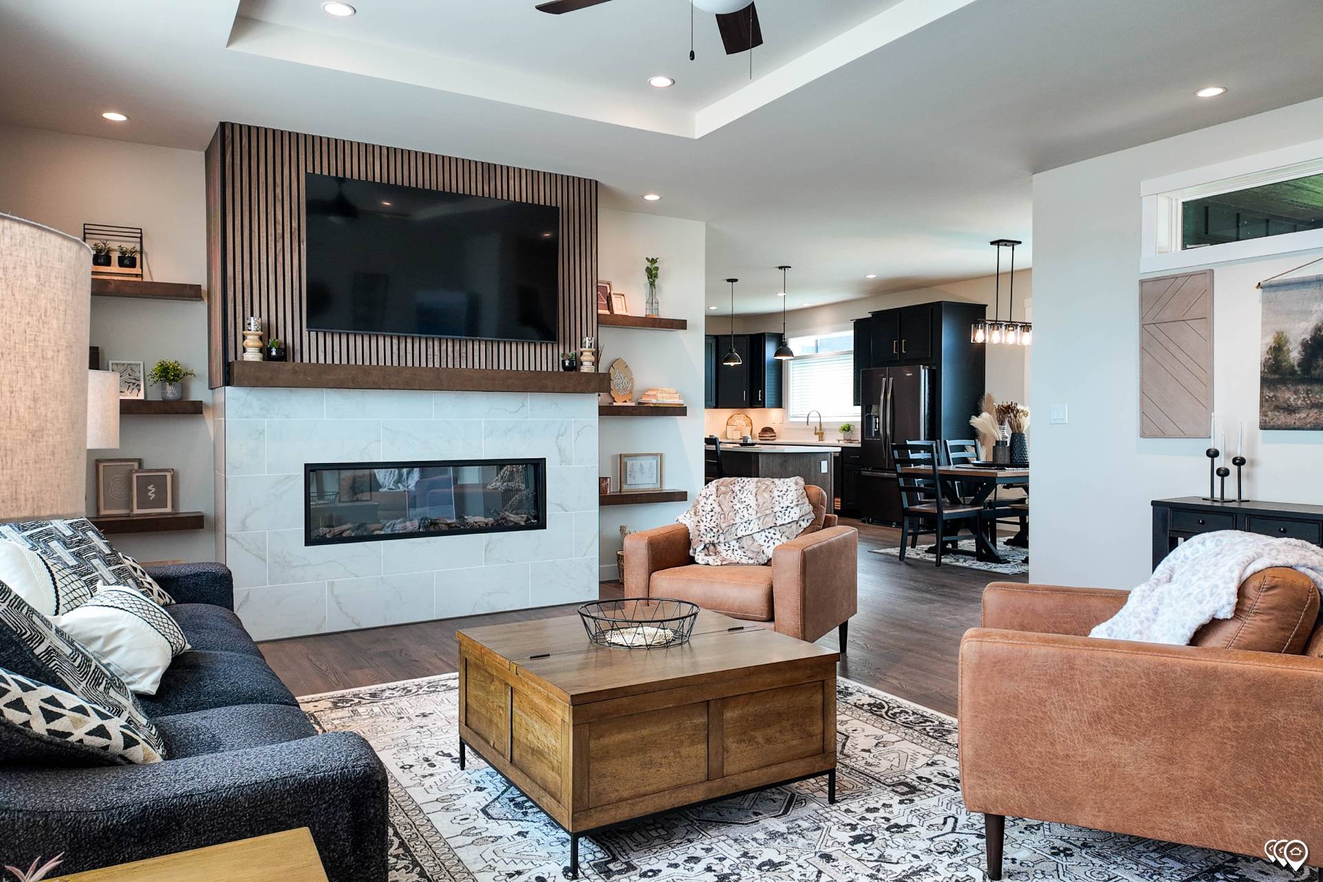 Modern living room with a cozy ambiance features a large TV above a sleek fireplace. A patterned rug, dark sofas, and tan chairs add warmth. Open kitchen and dining area with pendant lights in the background.