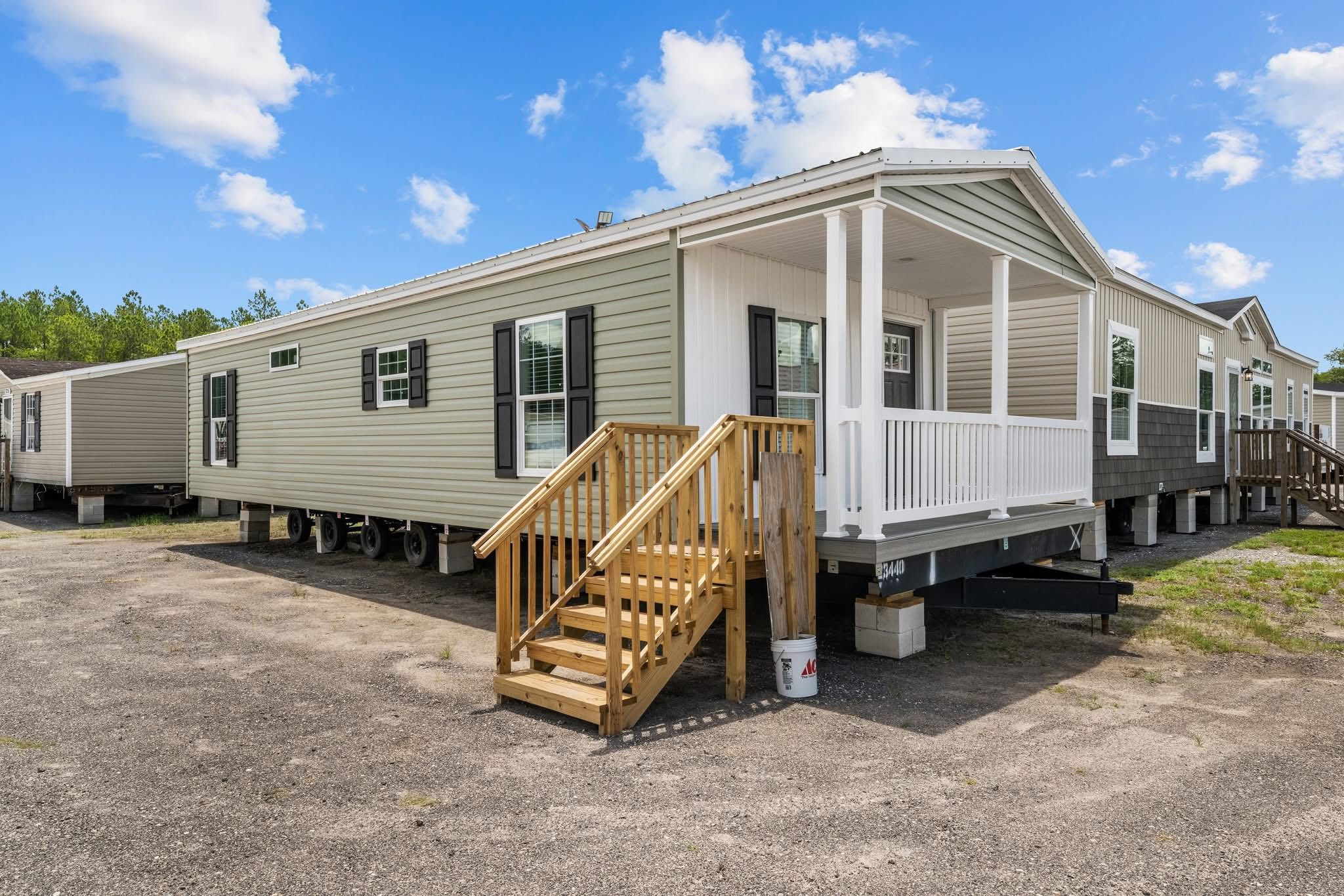 A row of mobile homes under a clear blue sky, with one in the foreground featuring beige siding, a small wooden porch, and black shutters.