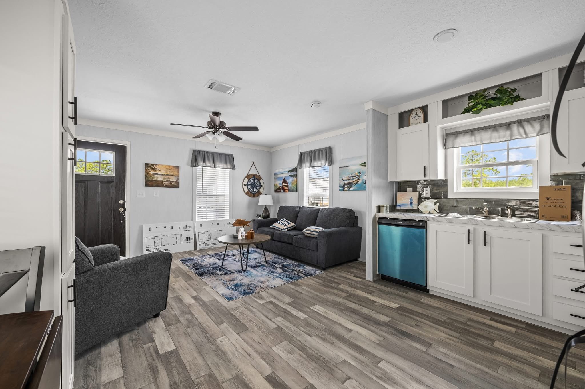 Modern living room with gray sofas, a round coffee table, and coastal decor. Open kitchen with white cabinets and stainless appliances. Bright, airy feel.