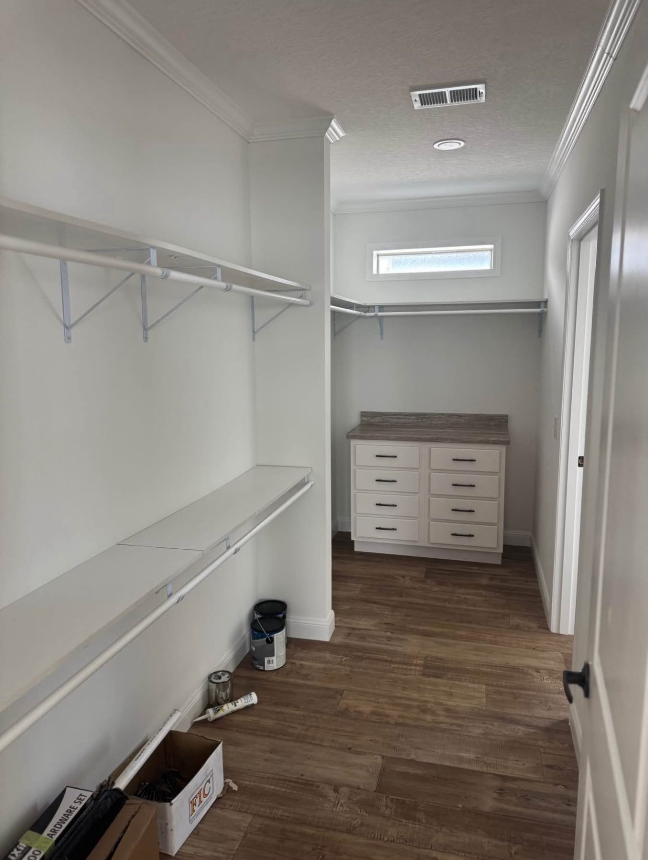 A spacious walk-in closet with white walls features wooden flooring, a dresser with multiple drawers, wire hanging racks, and a small high window.