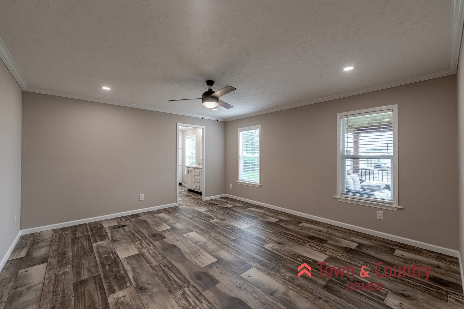 Spacious room with wood-patterned flooring, light gray walls, ceiling fan, and two windows. Doorway leads to a bright adjoining room. Calm and modern ambiance.