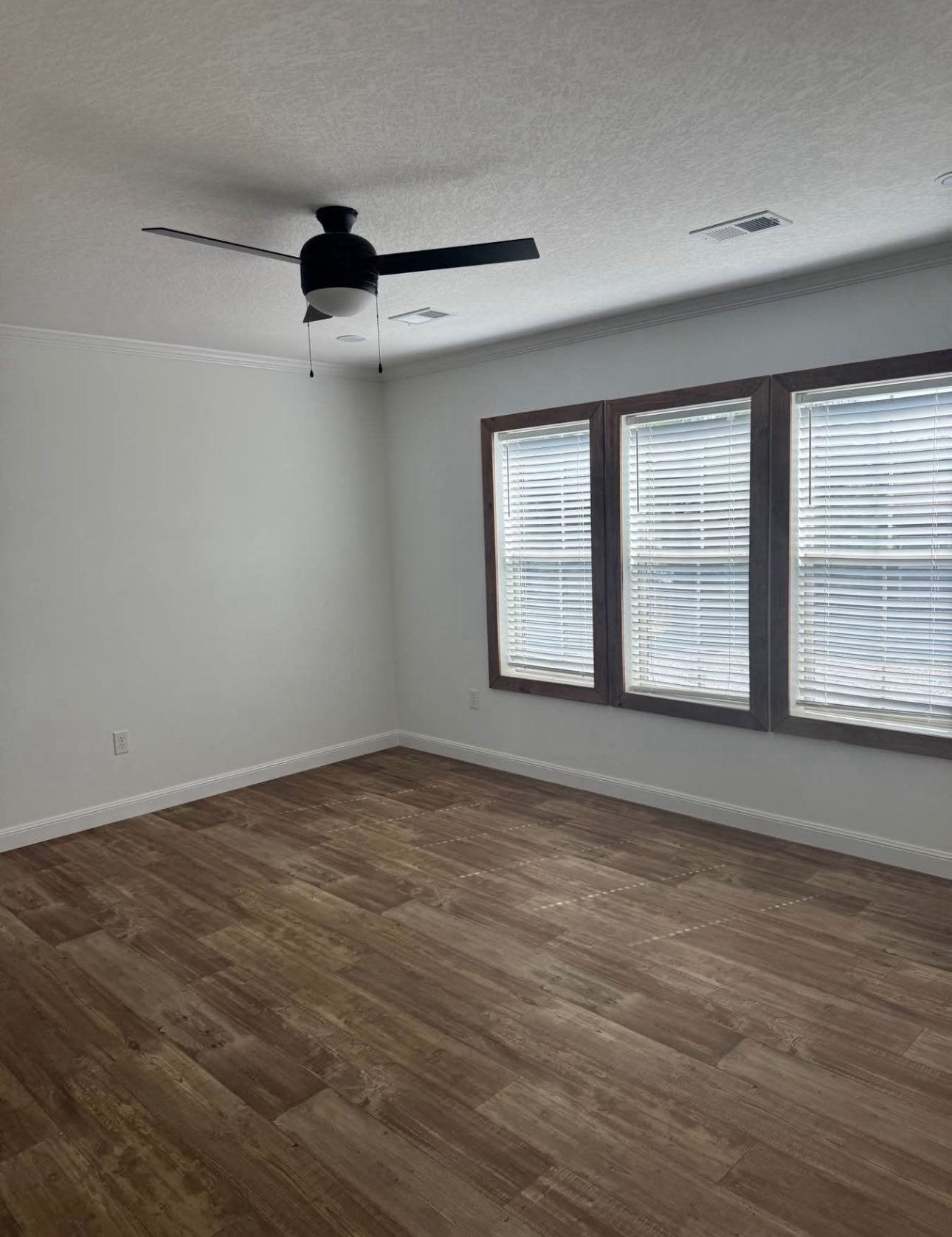Empty room with wooden floor, white walls, and three large windows with blinds on the right. A modern black ceiling fan is centered above. Calm and minimalist.