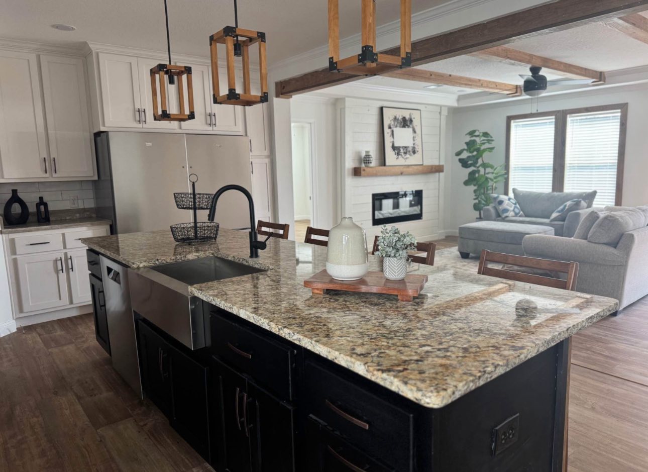 Modern kitchen with a large granite island, black base cabinets, and wood accents. Minimal decor, with pendant lights and adjacent cozy living area.