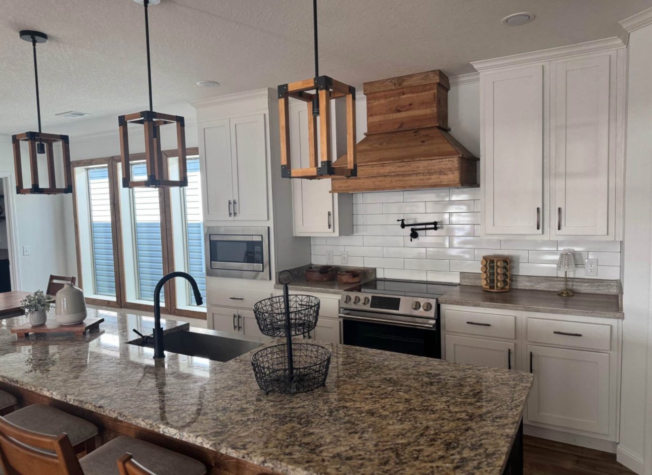 Modern kitchen with granite island, rustic wood range hood, and white cabinets. Three pendant lights hang above the island, creating a cozy atmosphere.