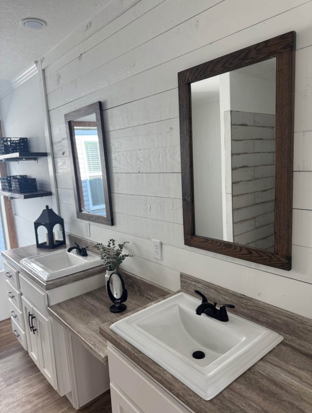 Double bathroom sinks with dark faucets, set in a rustic-style vanity. Two wooden-framed mirrors hang above, and shelves with black baskets are visible.