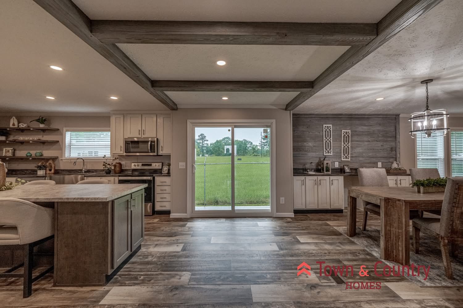 Spacious modern kitchen and dining area with wood floors and ceiling beams. Large window view of greenery. Cozy, inviting atmosphere.