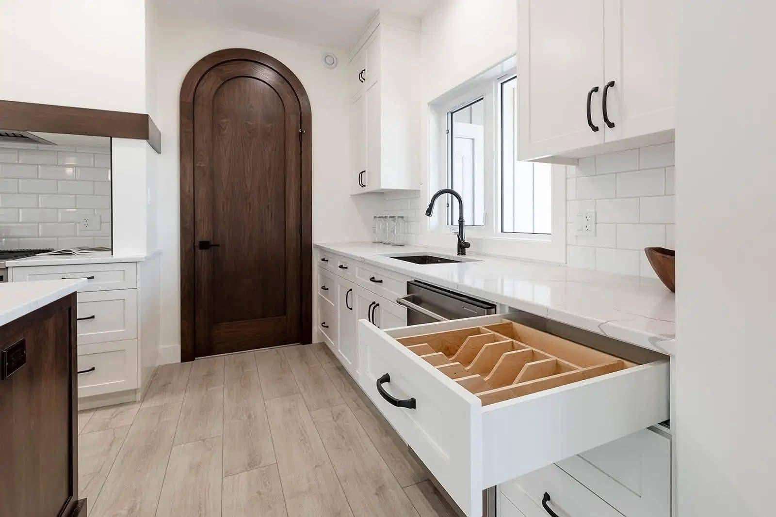 A modern kitchen with light wood flooring, white cabinets, and a drawer open revealing dividers. There is a dark wood arched door, white subway tiles, and a sleek black faucet.