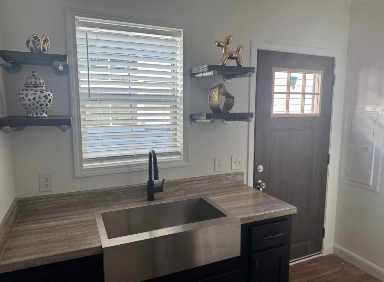 Modern kitchen with a sleek metal sink, dark faucet, and gray countertop. Shelves hold decorative items. A window and wooden door bring in natural light.