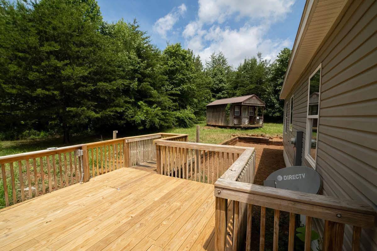 Wooden deck adjacent to a house with beige siding, overlooking a green forest. A wooden shed is in the yard. Clear skies add a serene mood.