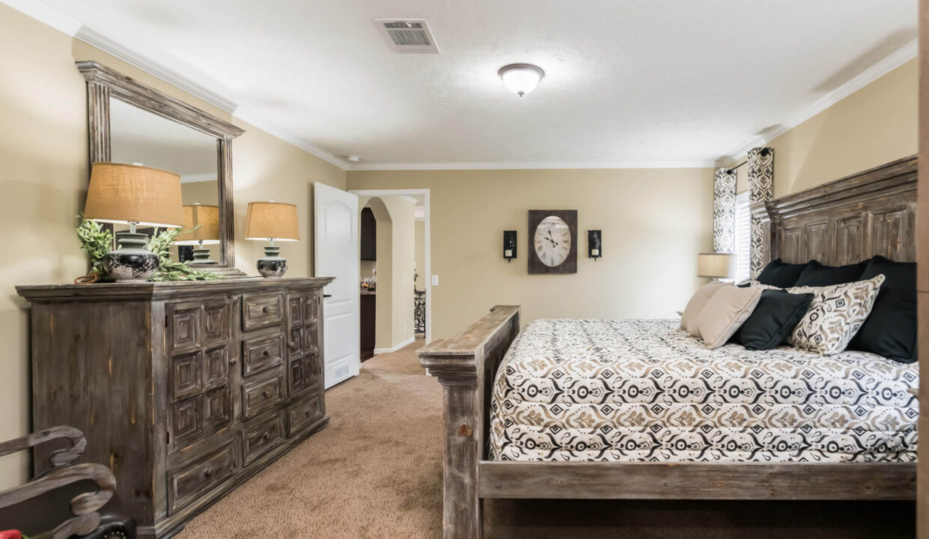 Cozy bedroom with a rustic wooden bed and dresser, beige walls, patterned bedding, and soft lighting. A clock and decorative pillows add charm.
