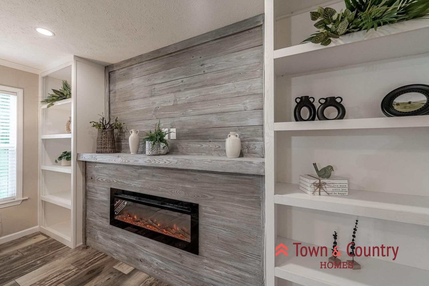 Cozy living room with a modern gray wooden accent wall featuring a linear electric fireplace. Built-in white shelves hold plants and decor. Bright, serene ambiance.