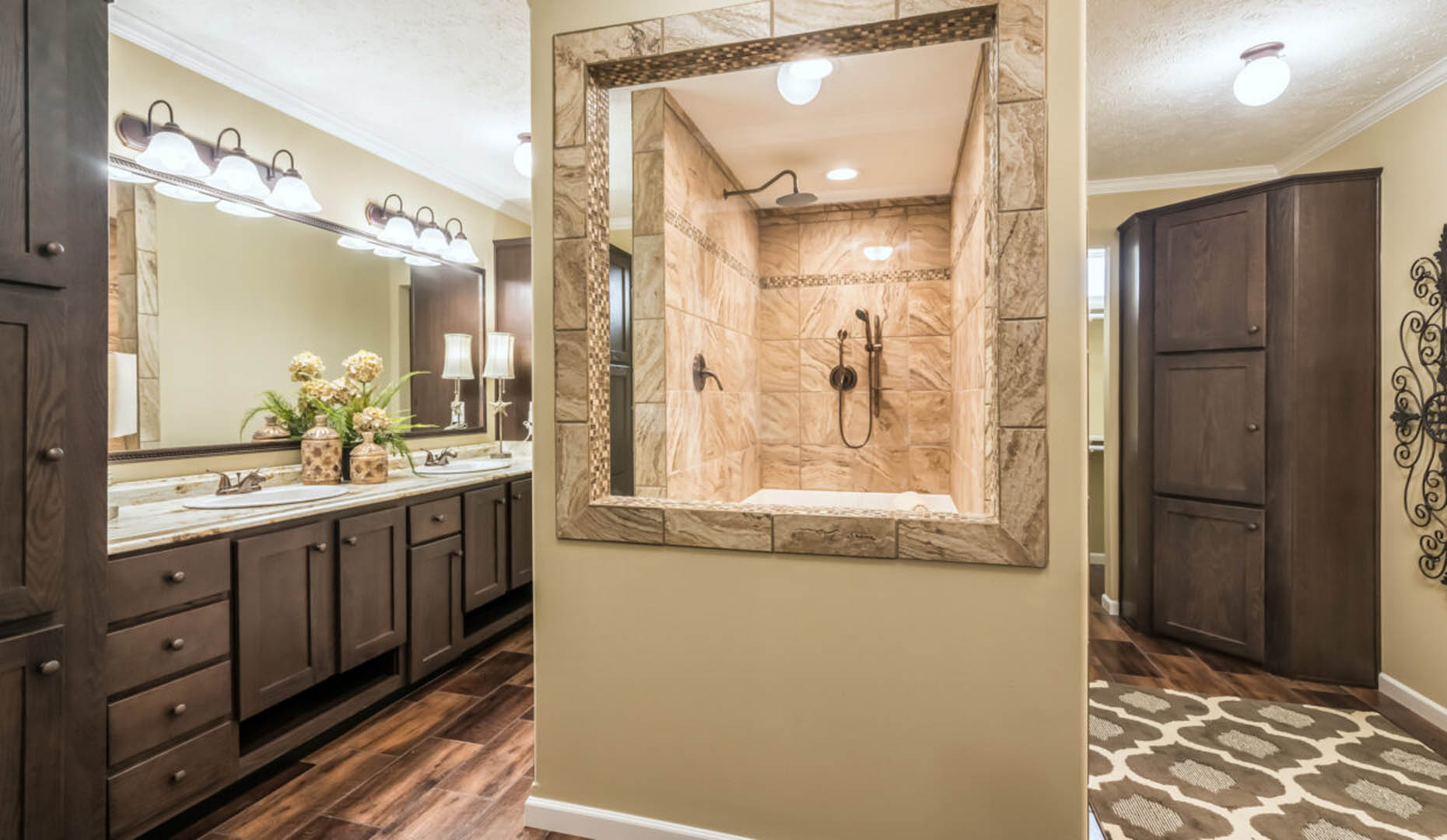 Elegant bathroom with dual sinks on a wooden vanity, decorative lamps, flowers, and a central tiled shower with earthy tones. Warm, inviting ambiance.