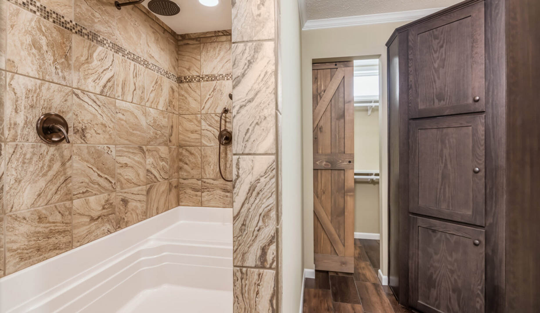Spacious bathroom with beige marble-style tiles, a white bathtub, and dark fixtures. A rustic wooden door leads to a closet. Warm, inviting atmosphere.