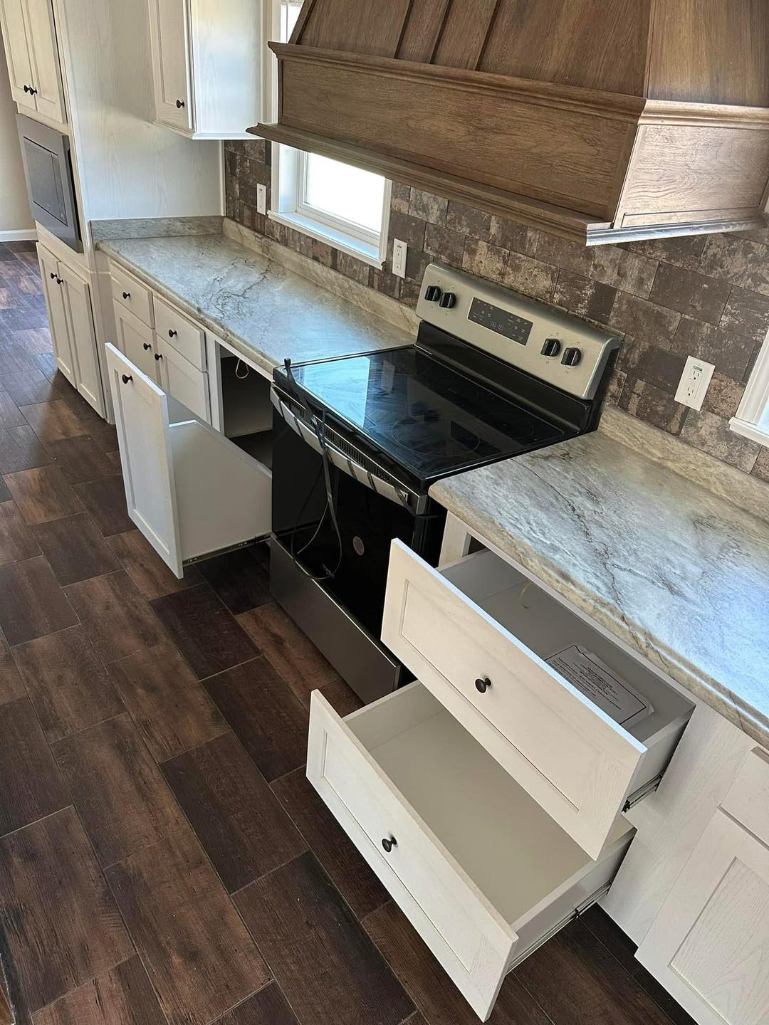 Spacious kitchen with dark wood floors, light countertops, and open white cabinets and drawers. A sleek black stove is centered under a wood hood.
