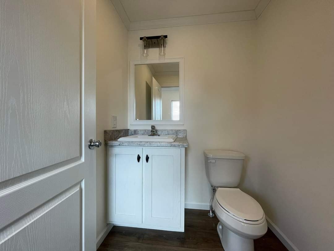 Small bathroom with a white vanity, mirror, and overhead light. Adjacent is a toilet. Space has beige walls and dark wood flooring, creating a simple, clean look.