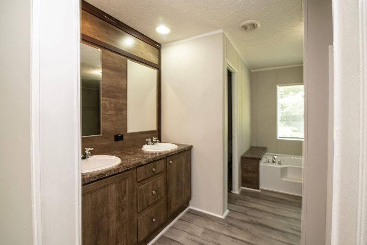 Modern bathroom with dual sinks and large wooden mirrors on the left. Gray flooring leads to a bathtub and window on the right, creating a bright, clean feel.
