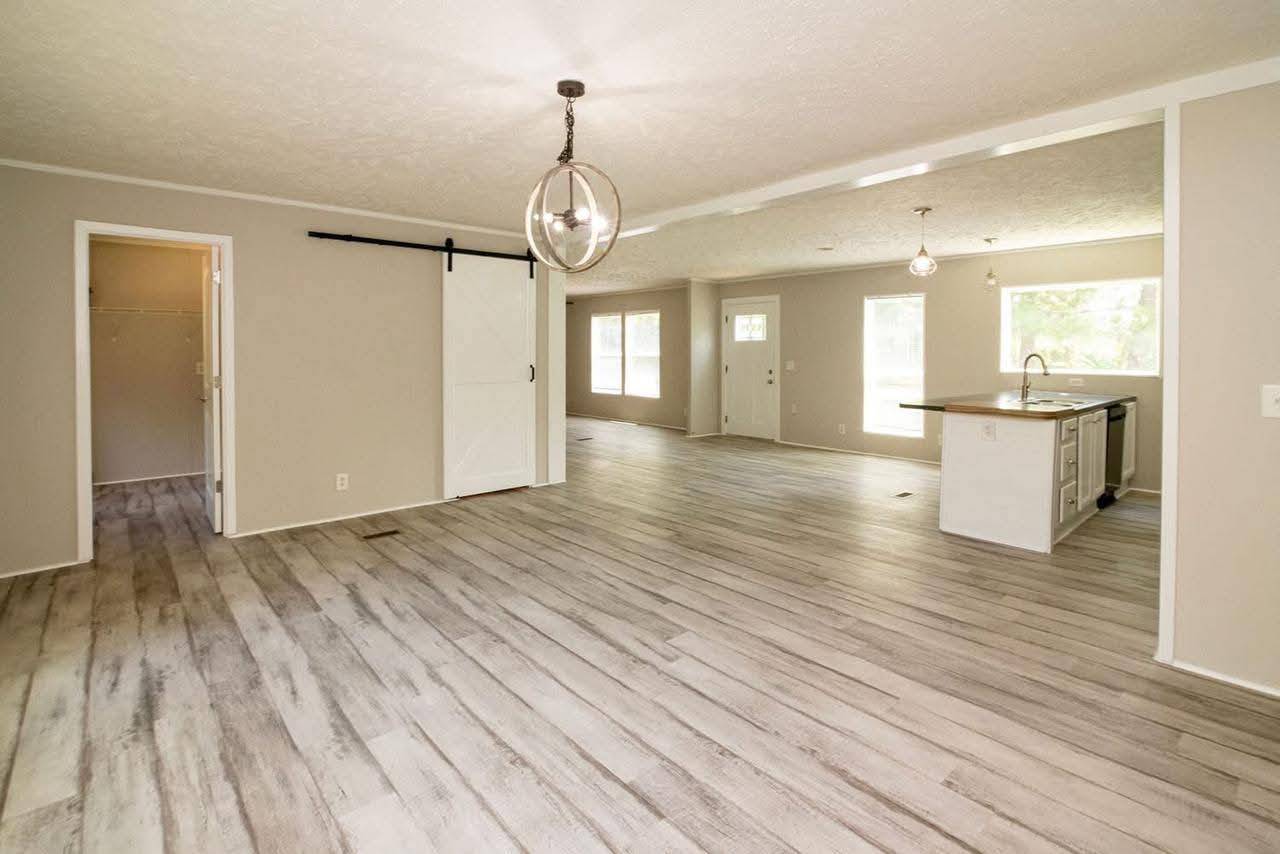 Spacious open floor plan with light wood flooring, a globe pendant light, a sliding barn door on the left, and a modern kitchen with island on the right.